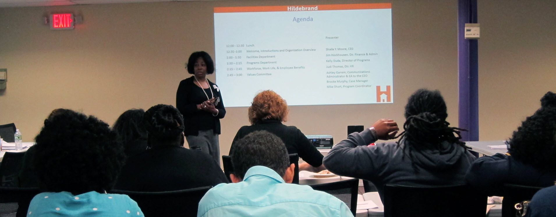 African-American profession woman conducting a workshop with powerpoints in a full classroom of adults.