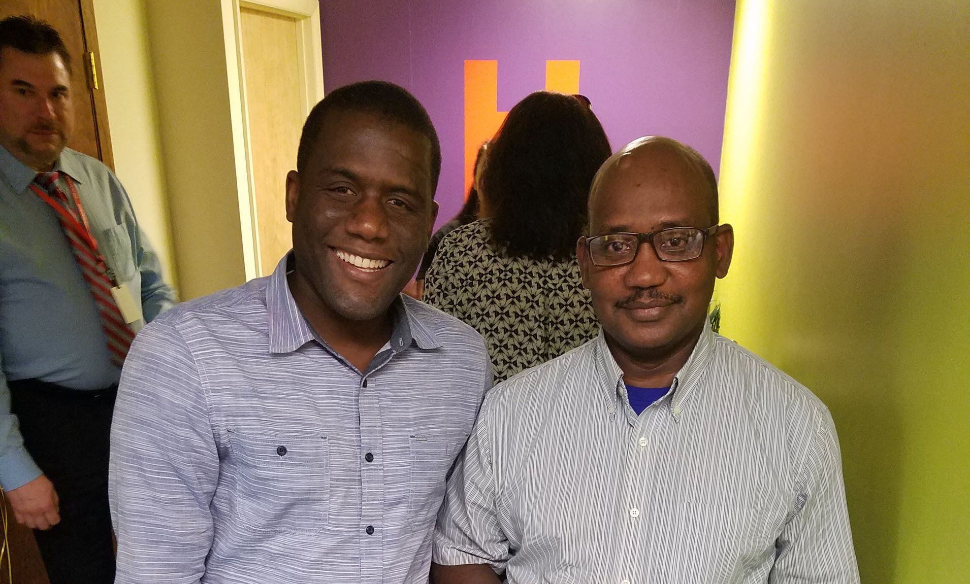 An African-American Man and South Asian-American man smiling for the camera with people and the Hildebrand logo visible in background.