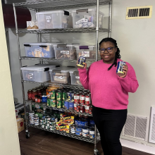 A Hildebrand staff member stands in front of the on-site food pantry holding cans of beans