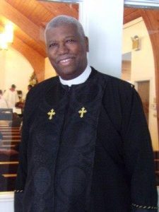 A man wearing pastor's robes smiles at the camera.